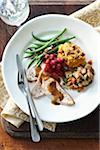 Overhead image of a holiday dinner plate containing turkey breast, gravy, stuffing, sweet potato, green beans, cranberries, walnuts and fresh rosemary. Cutlery, ice water, and a napkin are also present on a wooden cutting board on a table top.