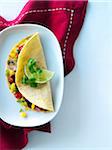 One vegetarian quesadilla in a yellow corn tortilla with cilantro, lime, mushrooms, corn, red pepper, melted cheese, and green onion. Mexican quesadilla is shown on a white cereamic plate with a red napkin on a tabletop.