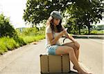 Teenage girl sitting on suitcase on the side of the road, looking at cell phone in summer, Germany