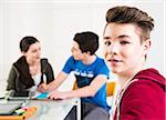 Teenage boy looking at camera, with teeange girl and teenage boy working on project together in background, studio shot