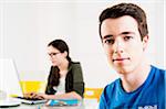 Teenage boy looking at camera, with teenage girl working on project using computer in background, studio shot