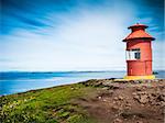 Little red lighthouse situated at the top of a cape over a sunny seascape. Related concept: navigation, help, aid, guide. Sykkisholmur, Iceland, Europe.