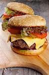 Two Ripe Hamburgers with Beef, Tomato, Lettuce, Pickled Cucumbers, Red Onion and Cheese into Sesame Buns closeup on Wooden Cutting Board
