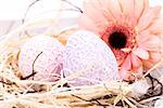 Beautiful pastel coloured Easter eggs in handcrafted intricate crocheted covers with a fresh pink Gerbera daisy in a clean straw nest for a traditional symbolic Easter celebration