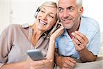 Cheerful mature man and woman with headphones and cellphone sitting on sofa at home