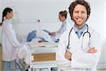 Portrait of male doctor standing arms crossed with colleagues examining patient in hospital