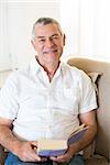 Portrait of smiling senior man holding book on sofa at home