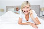 Portrait of a happy mature woman using cellphone while man using laptop in bed at home