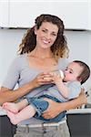 Portrait of happy mother feeding milk to baby boy from bottle in kitchen