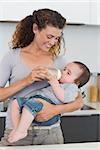 Beautiful woman feeding milk to baby from bottle in kitchen