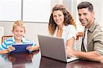 Happy parents sitting with son using tablet and laptop at home in kitchen