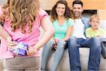 Little girl hiding present from her family behind back at home in living room