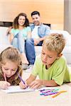 Siblings colouring on the rug with parents watching from sofa at home in living room