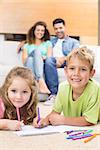 Happy siblings colouring on the rug with parents watching from sofa at home in living room