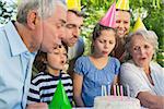 Cheerful extended family wearing party hats and blowing birthday cake in the park