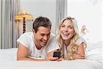 Cheerful relaxed casual young couple reading text message in bed at home