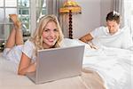 Smiling young woman using laptop while man reading book in background on bed at home