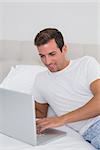 Smiling relaxed young man using laptop in bed at home