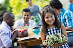 Adults and children around a table in a garden. A child holding a bowl of salad.
