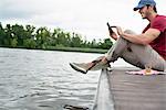 A man seated on a jetty by a lake, using a digital tablet.