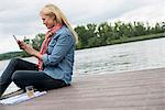 A woman sitting on a jetty by a lake, using a digital tablet.