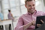 An open plan office in New York City.  A man in a pink shirt sitting smiling, using a digital tablet.