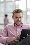 An open plan office in New York City.  A man in a pink shirt sitting smiling, using a digital tablet.