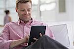 An open plan office in New York City.  A man in a pink shirt sitting smiling, using a digital tablet.  Wearing earphones.