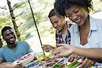 A family picnic meal in the shade of tall trees. Parents and children helping themselves to fresh fruits and vegetables.