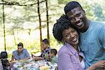 A family picnic meal in the shade of tall trees. A couple hugging each other.