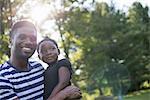 A man holding a small child in his arms, in the shade of trees on a hot summer day