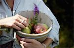 A woman holding a pottery bowl with fresh picked apples and a small foxglove flower.