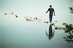 A man walking barefoot across stepping stones away from the shore of a lake.