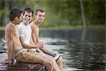 Three boys sitting on the jetty beside the water of a lake or river, wearing swimming trunks.