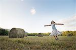 A field full of tall rounded hay bales, and a young girl dancing with her arms outstretched on the stubble field.