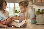 Two young girls sitting on a tabletop, holding a pot with a seedling and leaf shoot.