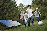 A group of boys running after a ball past solar panels in the woods.