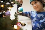 A young boy holding Christmas ornaments and placing them on the Christmas tree.