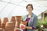 A woman wearing work gloves carrying terracotta plant pots in a large greenhouse on an organic farm.