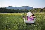 A young girl sitting in a traditional wooden Adirondack style chair in a field at evening light.