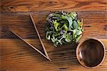 A small round polished wooden bowl and a clutch of organic mixed salad leaves, with wooden chopsticks.