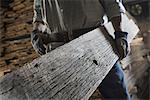 A heap of recycled reclaimed timber planks of wood. Environmentally responsible reclamation in a timber yard. A man carrying a large plank of mature weathered wood.