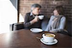 Two people sitting in a coffee shop. A man holding a white china of and drinking. Sitting beside a woman. A table with a large full cup of  cappuccino coffee.