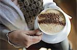 A person holding a full cup of frothy cappuccino coffee in a white china cup. Chocolate powder sprinkled in a pattern on the top. Coffee shop.