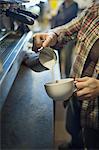 A person, barista, making coffee, and pouring frothed milk from a jug into a cup for a cappuccino. Coffee shop.