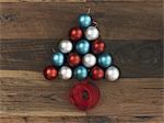 A collection of blue, red and silver ornaments arranged in a triangular shape on a wooden board. A Christmas tree shape with a red ribbon coiled at the base.