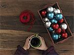 A collection of blue, red and silver ornaments and red ribbon in a box on a wooden board. A hand curled around a cup of coffee. Taking a break.