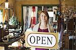 A woman standing in an antique store, holding an OPEN sign. Displays of goods all around her.