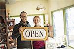 Two people standing in a store full of antique objects, a couple running a business. Holding a large sign saying OPEN.