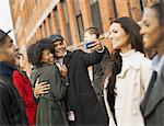 City life. A group of people on the go. A man holding out a camera phone and taking pictures of the group. Kissing a young woman. Men and women.